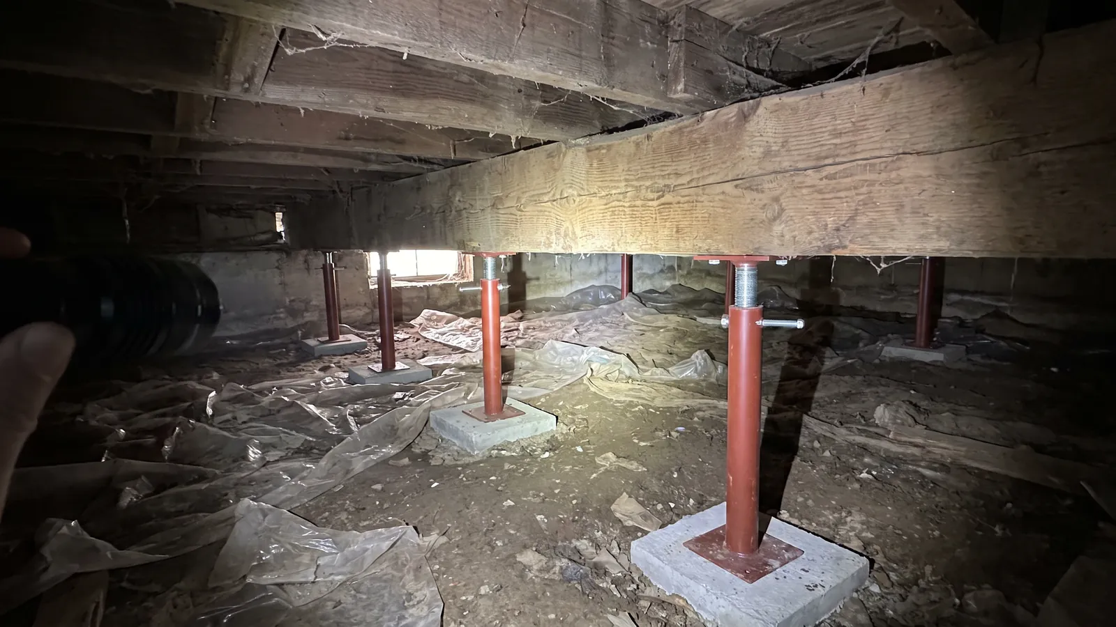 Crawl space under an older house with new adjustable steel support posts on concrete pads holding up a sagging wood girder